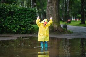 wenig Junge spielen im regnerisch Sommer- Park. Kind mit Regenschirm, wasserdicht Mantel und Stiefel Springen im Pfütze und Schlamm im das Regen. Kind Gehen im Sommer- Regen draussen Spaß durch irgendein Wetter. glücklich Kindheit foto