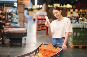 Frau beim Supermarkt halten ein voll Einkaufen Wagen und ein Einkaufen aufführen foto