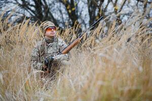 ein männlich Jäger mit ein Gewehr während Sitzung nimmt Ziel beim ein Wald. das Konzept von ein erfolgreich Jagd, ein erfahren Jäger. Jagd das Herbst Jahreszeit. das Jäger hat ein Gewehr und ein Jagd Uniform foto