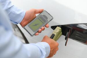 glücklich Mann mit Clever Telefon und Laden Auto beim Fahrzeug Laden Bahnhof foto