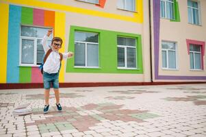 zurück zu Schule. glücklich lächelnd Junge im Brille ist gehen zu Schule zum das zuerst Zeit. Kind mit Rucksack und Buch draußen. Anfang von Unterricht. zuerst Tag von fallen foto