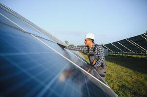 Porträt von jung indisch Mann Techniker tragen Weiß schwer Hut Stehen in der Nähe von Solar- Paneele gegen Blau Himmel. industriell Arbeiter Solar- System Installation foto