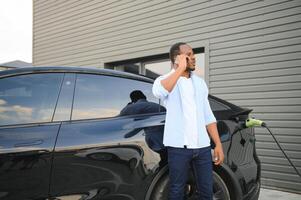 glücklich jung Mann mit Handy, Mobiltelefon Telefon Laden Auto beim elektrisch Fahrzeug Laden Bahnhof foto