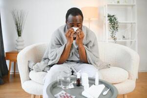 Porträt von ein krank jung afrikanisch amerikanisch Mann eingewickelt im ein Decke auf das Sofa. Kerl mit saisonal Grippe oder kalt Gefühl ungesund mit Grippe und flüssig Nase beim Zuhause foto