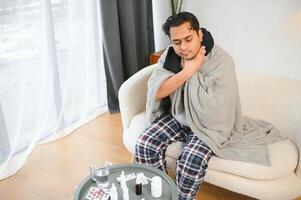 jung Muslim Mann Leiden von ein wund Kehle, Sitzung auf das Sofa beim Zuhause und Grimassen, halten seine Kehle. Bedürfnisse medizinisch Behandlung foto