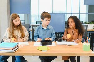 gemischtrassig Schulkinder haben Mittagessen beim das Schreibtisch während ein brechen im Schule foto