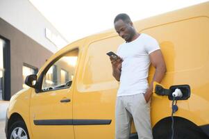 beiläufig afrikanisch Mann mit Smartphone in der Nähe von elektrisch Auto warten zum das Fertig von das Batterie Laden Verfahren. foto