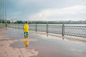 wenig Junge spielen im regnerisch Sommer- Park. Kind mit Regenschirm, wasserdicht Mantel und Stiefel Springen im Pfütze und Schlamm im das Regen. Kind Gehen im Sommer- Regen draussen Spaß durch irgendein Wetter. glücklich Kindheit. foto