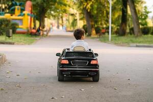 süß Junge im Reiten ein schwarz elektrisch Auto im das Park. komisch Junge Fahrten auf ein Spielzeug elektrisch Wagen. Kopieren Raum. foto