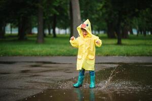 wenig Junge spielen im regnerisch Sommer- Park. Kind mit Regenschirm, wasserdicht Mantel und Stiefel Springen im Pfütze und Schlamm im das Regen. Kind Gehen im Sommer- Regen draussen Spaß durch irgendein Wetter. glücklich Kindheit. foto