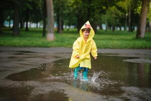 wenig Junge spielen im regnerisch Sommer- Park. Kind mit Regenschirm, wasserdicht Mantel und Stiefel Springen im Pfütze und Schlamm im das Regen. Kind Gehen im Sommer- Regen draussen Spaß durch irgendein Wetter. glücklich Kindheit. foto