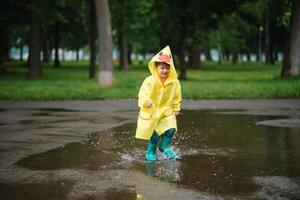 wenig Junge spielen im regnerisch Sommer- Park. Kind mit Regenschirm, wasserdicht Mantel und Stiefel Springen im Pfütze und Schlamm im das Regen. Kind Gehen im Sommer- Regen draussen Spaß durch irgendein Wetter. glücklich Kindheit. foto