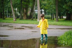 wenig Junge spielen im regnerisch Sommer- Park. Kind mit Regenschirm, wasserdicht Mantel und Stiefel Springen im Pfütze und Schlamm im das Regen. Kind Gehen im Sommer- Regen draussen Spaß durch irgendein Wetter. glücklich Kindheit. foto