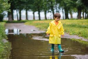 wenig Junge spielen im regnerisch Sommer- Park. Kind mit Regenschirm, wasserdicht Mantel und Stiefel Springen im Pfütze und Schlamm im das Regen. Kind Gehen im Sommer- Regen draussen Spaß durch irgendein Wetter. glücklich Kindheit foto