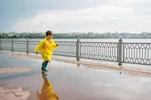 wenig Junge spielen im regnerisch Sommer- Park. Kind mit Regenschirm, wasserdicht Mantel und Stiefel Springen im Pfütze und Schlamm im das Regen. Kind Gehen im Sommer- Regen draussen Spaß durch irgendein Wetter. glücklich Kindheit. foto