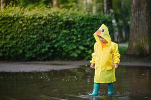 wenig Junge spielen im regnerisch Sommer- Park. Kind mit Regenschirm, wasserdicht Mantel und Stiefel Springen im Pfütze und Schlamm im das Regen. Kind Gehen im Sommer- Regen draussen Spaß durch irgendein Wetter. glücklich Kindheit foto