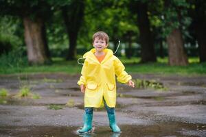 wenig Junge spielen im regnerisch Sommer- Park. Kind mit Regenschirm, wasserdicht Mantel und Stiefel Springen im Pfütze und Schlamm im das Regen. Kind Gehen im Sommer- Regen draussen Spaß durch irgendein Wetter. glücklich Kindheit foto