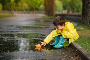 Kind spielen mit Spielzeug Boot im Pfütze. Kind abspielen draussen durch Regen. fallen regnerisch Wetter draußen Aktivität zum jung Kinder. Kind Springen im schlammig Pfützen. wasserdicht Jacke und Stiefel zum Baby. Kindheit foto