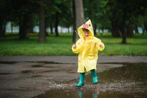 wenig Junge spielen im regnerisch Sommer- Park. Kind mit Regenschirm, wasserdicht Mantel und Stiefel Springen im Pfütze und Schlamm im das Regen. Kind Gehen im Sommer- Regen draussen Spaß durch irgendein Wetter. glücklich Kindheit. foto