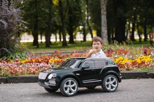 süß Junge im Reiten ein schwarz elektrisch Auto im das Park. komisch Junge Fahrten auf ein Spielzeug elektrisch Wagen. Kopieren Raum. foto