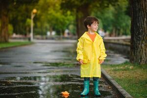 Kind spielen mit Spielzeug Boot im Pfütze. Kind abspielen draussen durch Regen. fallen regnerisch Wetter draußen Aktivität zum jung Kinder. Kind Springen im schlammig Pfützen. wasserdicht Jacke und Stiefel zum Baby. Kindheit foto