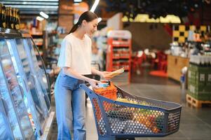 lächelnd glücklich Frau genießen Einkaufen beim das Supermarkt, sie ist gelehnt auf ein voll Wagen foto