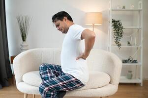 ein jung indisch Mann ist Leiden von stark Schmerz. er sitzt beim Zuhause auf das Sofa und hält seine Hand hinter seine zurück foto