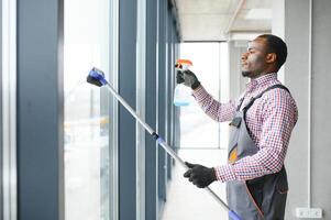 Arbeiter von Reinigung Organisation vorsichtig und vorsichtig reibt groß Fenster von das Büro Raum. ein ernst Afroamerikaner im Blau Overall Tücher das doppelt verglast Fenster im das Büro foto