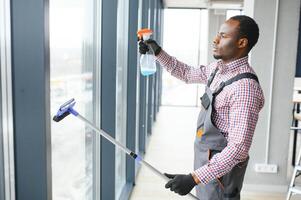 afrikanisch männlich Fachmann Reinigung Bedienung Arbeiter im Overall reinigt das Fenster und Geschäft Fenster von ein Geschäft mit Besondere Ausrüstung foto