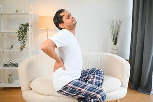 ein jung indisch Mann ist Leiden von stark Schmerz. er sitzt beim Zuhause auf das Sofa und hält seine Hand hinter seine zurück foto