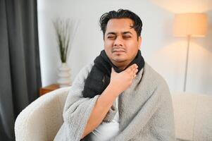 jung Muslim Mann Leiden von ein wund Kehle, Sitzung auf das Sofa beim Zuhause und Grimassen, halten seine Kehle. Bedürfnisse medizinisch Behandlung foto