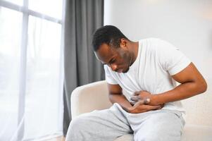 afrikanisch amerikanisch Kerl haben Bauch schmerzen nach Essen berühren Schmerz Bauch Leiden von Schmerzen Sitzung auf Sofa beim heim. foto