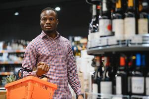 afrikanisch amerikanisch Mann halten Flasche von Wein und suchen beim es während Stehen im ein Wein Geschäft foto