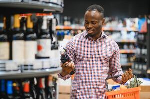 afrikanisch amerikanisch Mann halten Flasche von Wein und suchen beim es während Stehen im ein Wein Geschäft foto