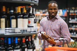 afrikanisch amerikanisch Mann halten Flasche von Wein und suchen beim es während Stehen im ein Wein Geschäft foto