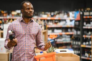 afrikanisch amerikanisch Mann halten Flasche von Wein und suchen beim es während Stehen im ein Wein Geschäft foto