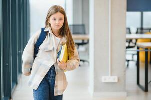 ziemlich blond Schule Mädchen halten viele bunt Anmerkungen und Bücher. klug Teen Mädchen lächelnd beim Kamera, Stehen auf Gang von International Schule foto