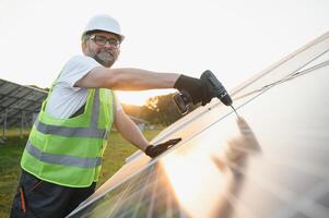 Porträt von lächelnd zuversichtlich Ingenieur Techniker mit elektrisch Schraubendreher, Stehen im Vorderseite von unvollendet hoch Außen Solar- Panel Foto voltaic System