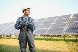 Porträt von jung indisch Mann Techniker tragen Weiß schwer Hut Stehen in der Nähe von Solar- Paneele gegen Blau Himmel. industriell Arbeiter Solar- System Installation foto