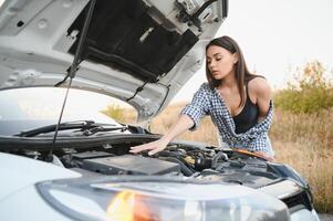 schön schlank Mädchen im Hemd und kurze Hose sieht aus im öffnen Auto Kapuze auf ein Straße foto