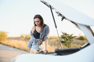 schön schlank Mädchen im Hemd und kurze Hose sieht aus im öffnen Auto Kapuze auf ein Straße foto