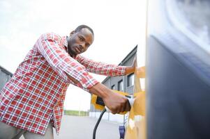 afrikanisch Mann halten aufladen Kabel im auf Hand Stehen in der Nähe von elektrisch Auto foto