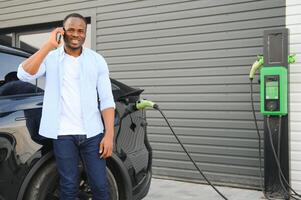 ein afrikanisch Mann ist Stehen in der Nähe von ein elektrisch Auto, warten zum es zu aufladen beim ein Laden Bahnhof und mit das Telefon. foto