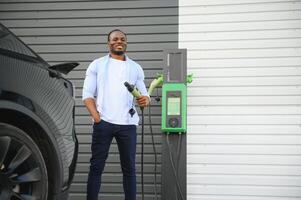 ernst afrikanisch Mann halten aufladen Kabel im auf Hand, Stehen in der Nähe von Luxus elektrisch Wagen. foto