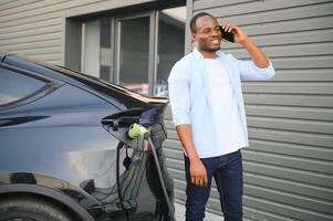 afrikanisch Mann Stehen in der Nähe von elektrisch Auto mit Laden Kabel im Stecker. Öko freundlich Fahrzeug Laden auf Bahnhof. Lebensstile Konzept foto