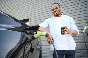 afrikanisch Mann Stehen in der Nähe von elektrisch Auto mit Laden Kabel im Stecker. Öko freundlich Fahrzeug Laden auf Bahnhof. Lebensstile Konzept foto