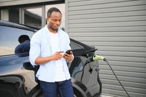 ein afrikanisch Mann ist Stehen in der Nähe von ein elektrisch Auto, warten zum es zu aufladen beim ein Laden Bahnhof und mit das Telefon. foto