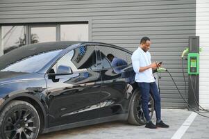 afrikanisch Mann Stehen in der Nähe von elektrisch Auto mit Laden Kabel im Stecker. Öko freundlich Fahrzeug Laden auf Bahnhof. Lebensstile Konzept foto