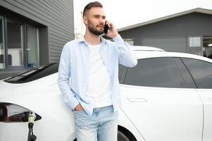 glücklich Mann mit Clever Telefon und Laden Auto beim Fahrzeug Laden Bahnhof foto