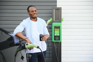 afrikanisch Mann Stehen in der Nähe von elektrisch Auto mit Laden Kabel im Stecker. Öko freundlich Fahrzeug Laden auf Bahnhof. Lebensstile Konzept foto